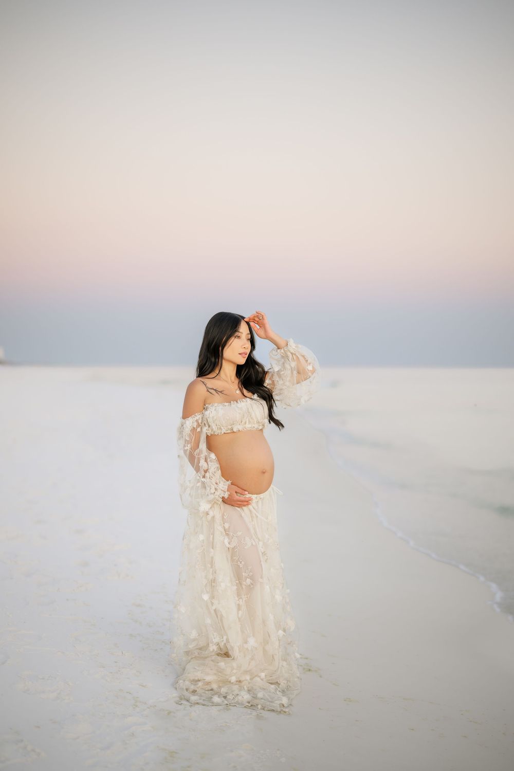 Expectant mother in flowing lace gown poses gracefully on white salt flats during sunset maternity photoshoot.