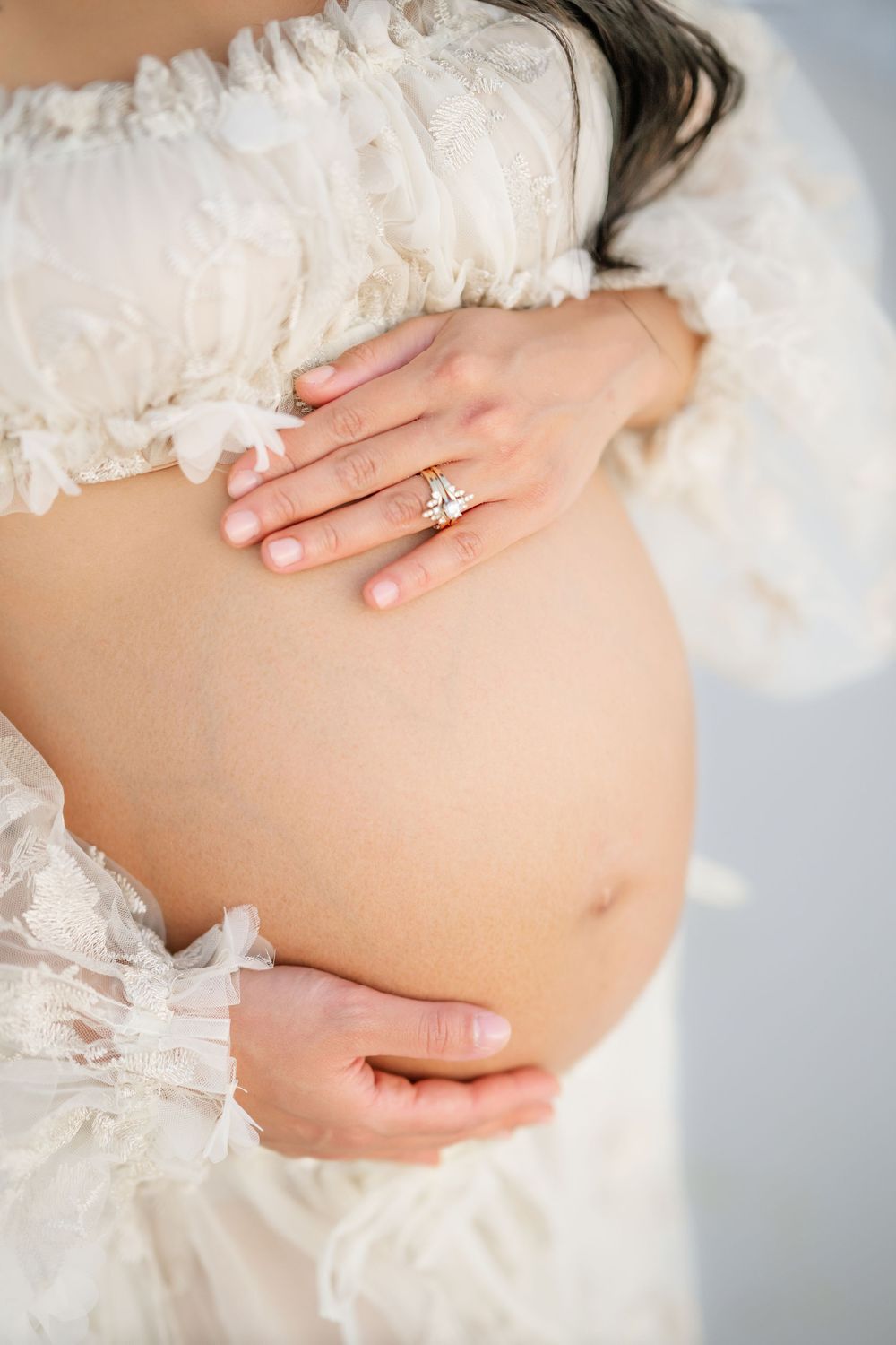 Close-up of pregnant belly with hands wearing engagement ring and white lace maternity dress details.