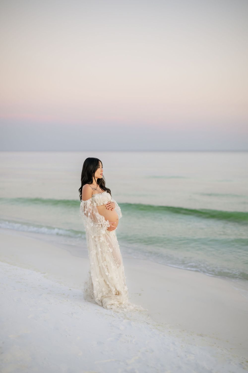 Expectant woman in flowing lace dress stands on white sandy beach at sunset Panama City beach maternity photoshoot, best photographer