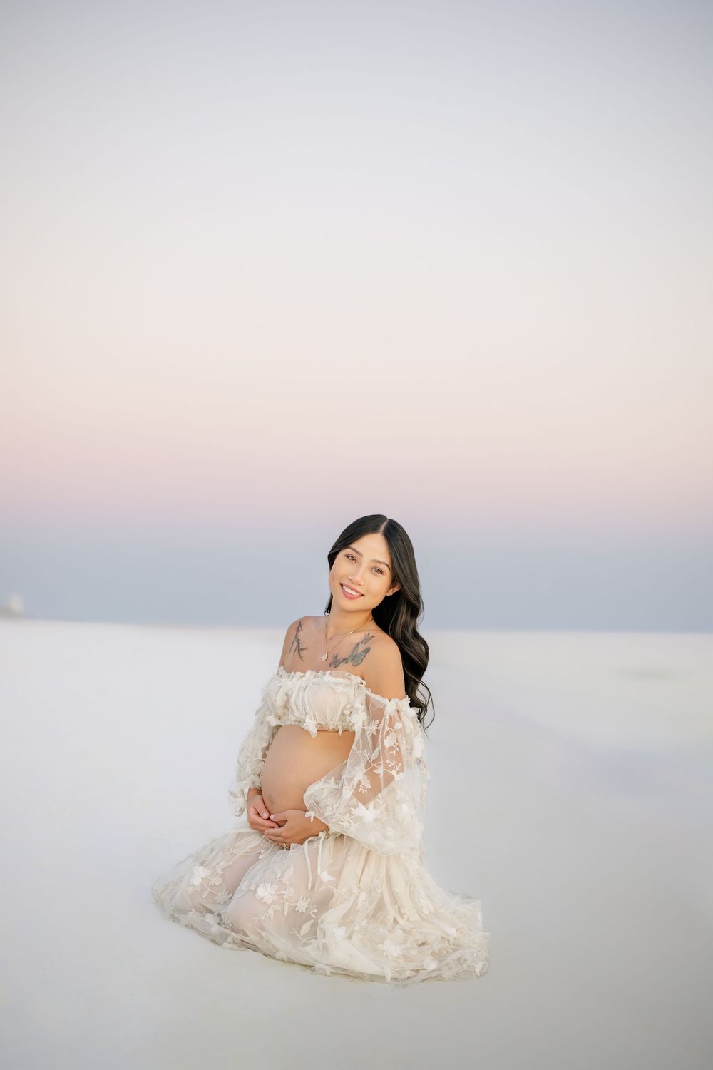 Woman in delicate lace gown kneels on white sand during sunset maternity photoshoot.Panama City beach maternity photoshoot, best photographe