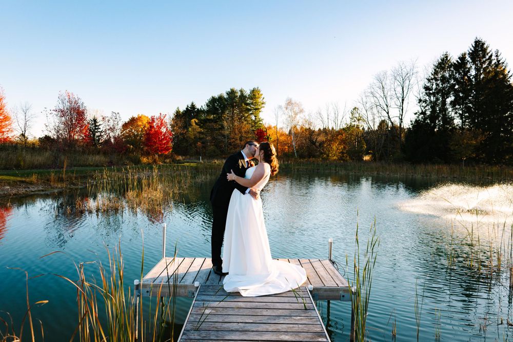 A couple embraces on a wooden dock overlooking a tranquil pond during sunset with autumn foliage in the background.