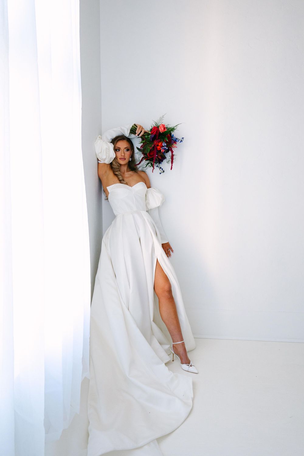 Elegant off-shoulder wedding gown with long sleeves and high slit shows alongside vibrant red and pink bouquet against white wall.