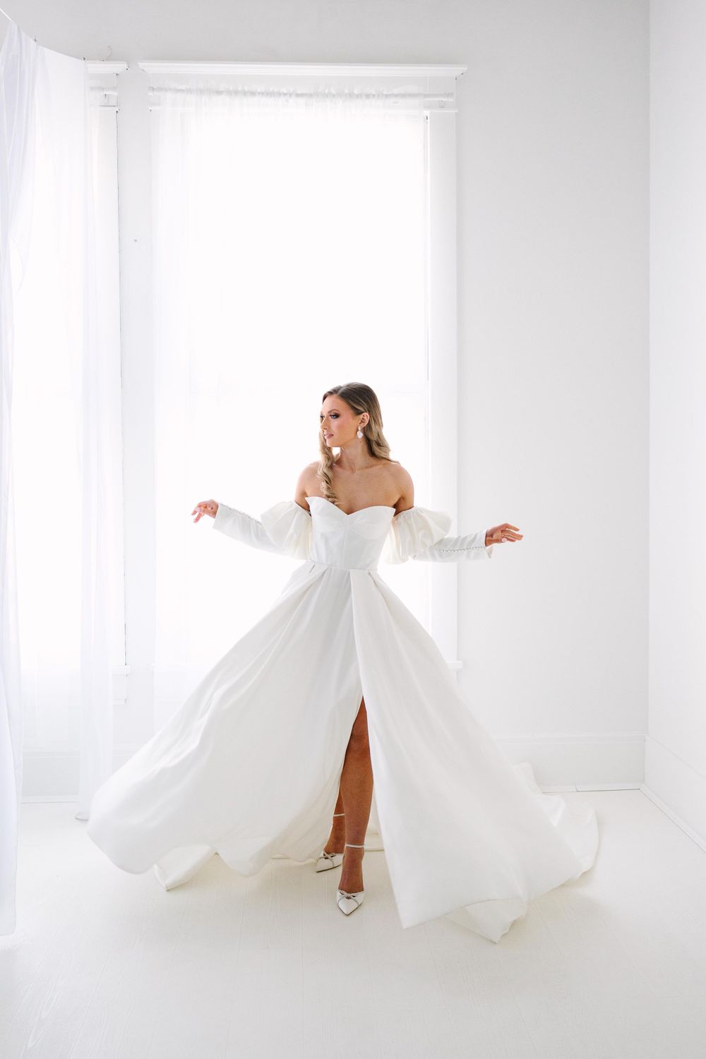 A bride in an elegant off-shoulder white gown with long sleeves twirls in a bright minimalist room.