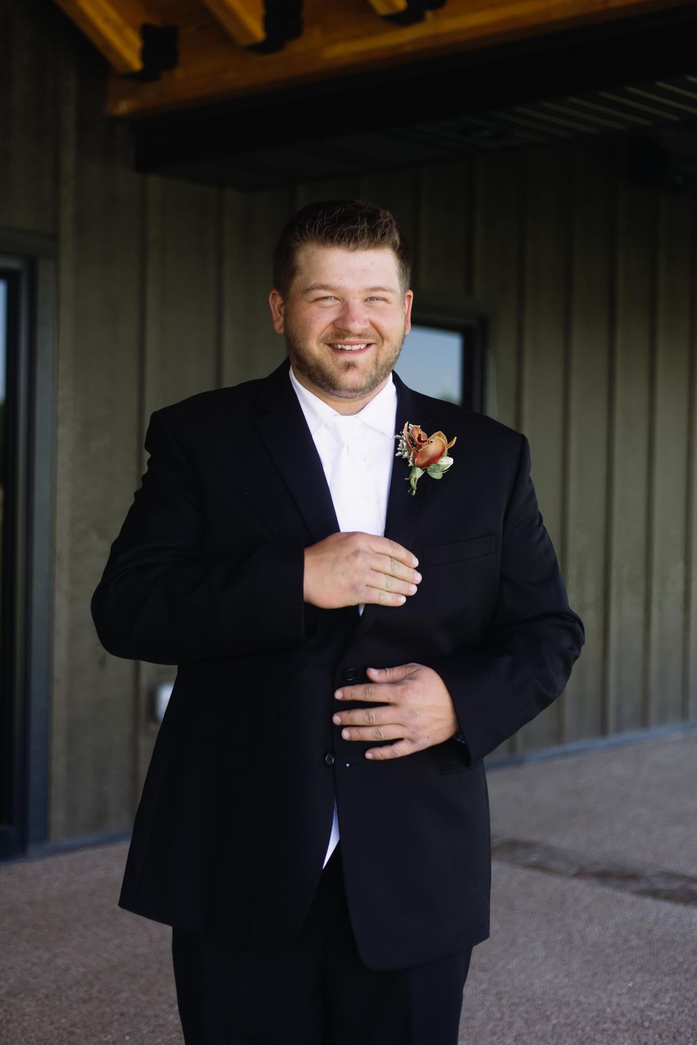 A groom in a black tuxedo smiles while wearing a floral boutonniere.