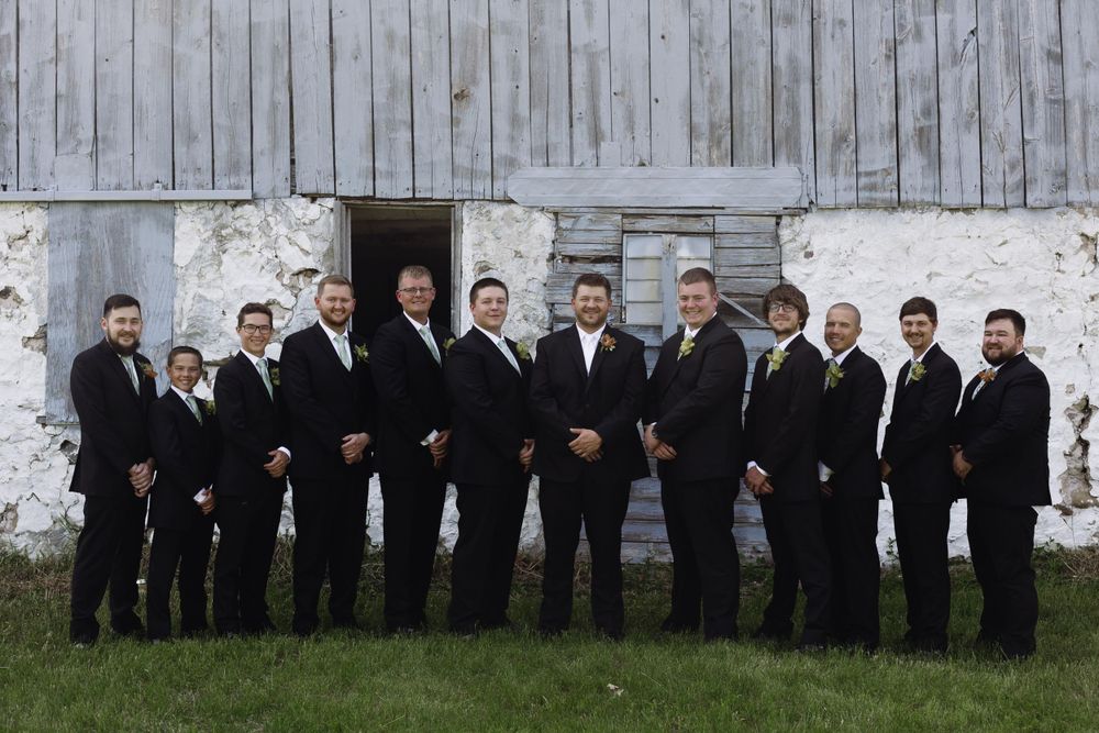 Groomsmen in black suits line up against white barn wall for formal wedding photo.