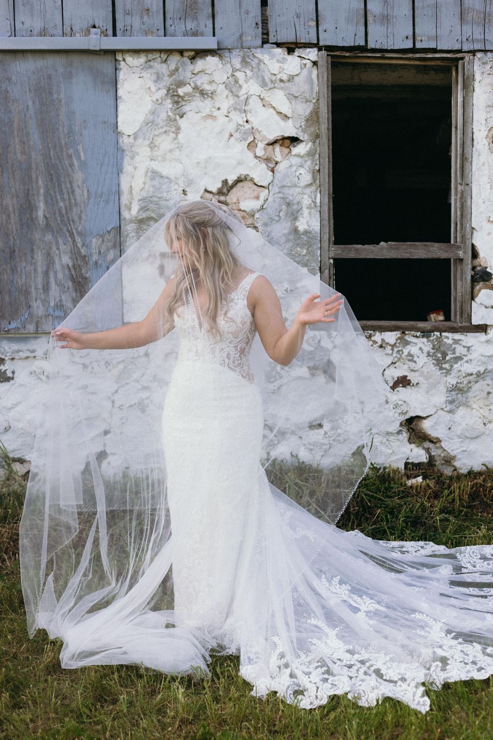 Bride in lace wedding gown with flowing veil poses against weathered building.