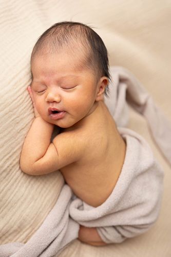 A sleeping newborn baby in a soft gray onesie rests peacefully on cream-colored bedding.