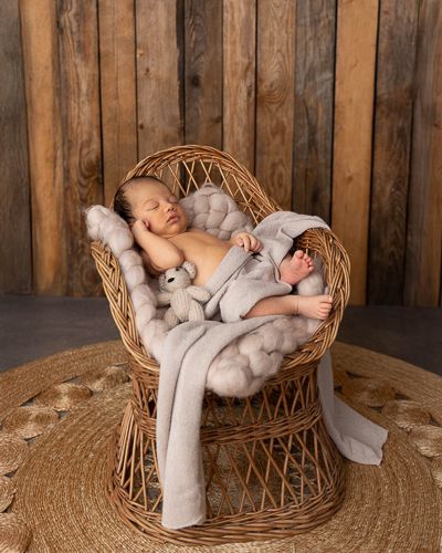 A sleeping baby rests peacefully in a woven rattan chair against a rustic wooden wall background.