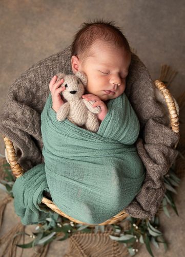A sleeping newborn swaddled in mint green muslin wrap on a grey blanket with eucalyptus leaves and natural decor.