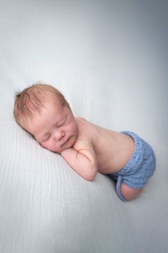 A sleeping newborn baby wearing blue knit pants lies peacefully on a white textured surface.