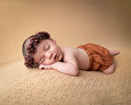 A sleeping baby in a rust-colored romper lies on a textured beige backdrop in a creative newborn photography setup.