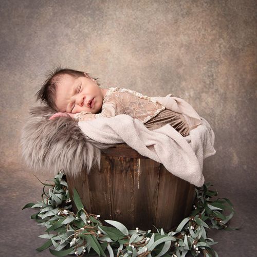 Nyfødt baby I en rustikk trebøtte omgitt av eukalyptusblader på brun bakgrunn.Nyfødtfotografering i studio på Fjerdingby på Romerike