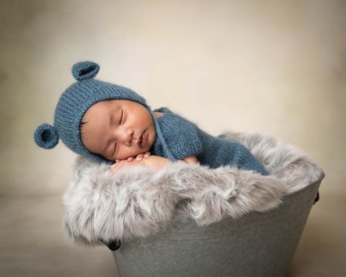 Sovende baby med blått antrekk med bjørneører på luen. Ligger i en grå bøtte med grått fluffy  skinn. Nyfødtfotografering i studio på Fjer