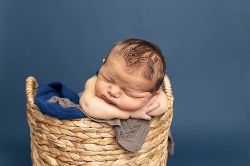 A sleeping newborn cradled in a round woven basket on textured fabric with blue background.