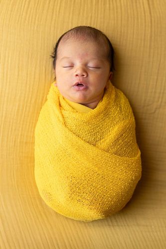 A sleeping newborn baby swaddled in a bright yellow knit wrap against a cream-colored background.