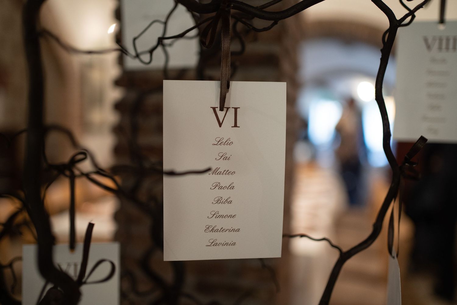 A wedding seating chart table number VI hangs from dark branches with blurred background decor.