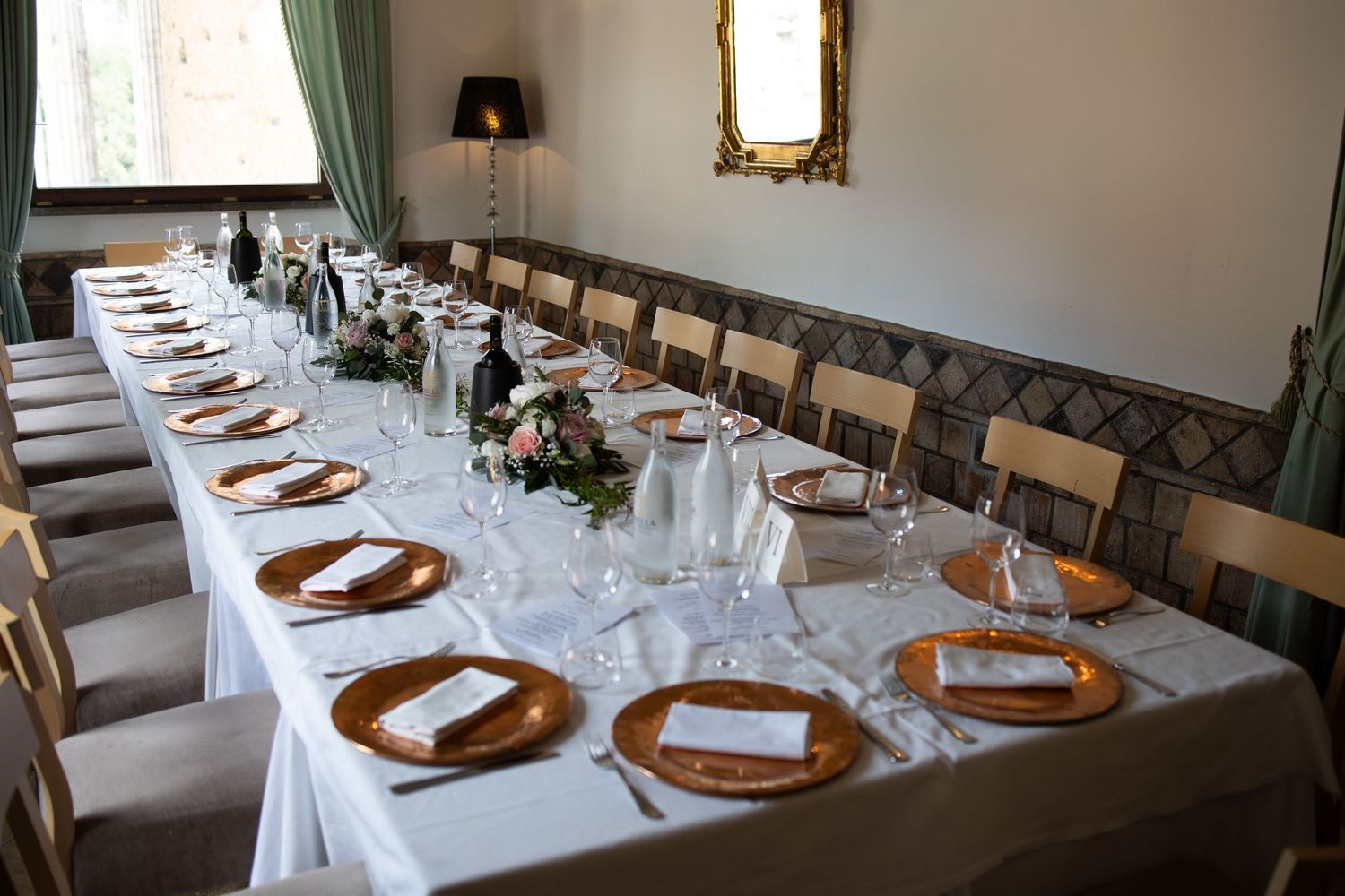 Elegant banquet hall with long dining tables set with white linens, glassware and wooden chairs in a formal setting.