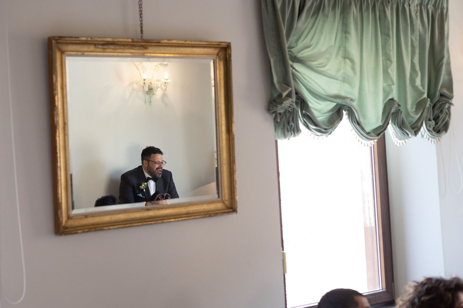 A reflection in a gold-framed mirror shows someone in formal attire seated against a light wall with sage green curtains.