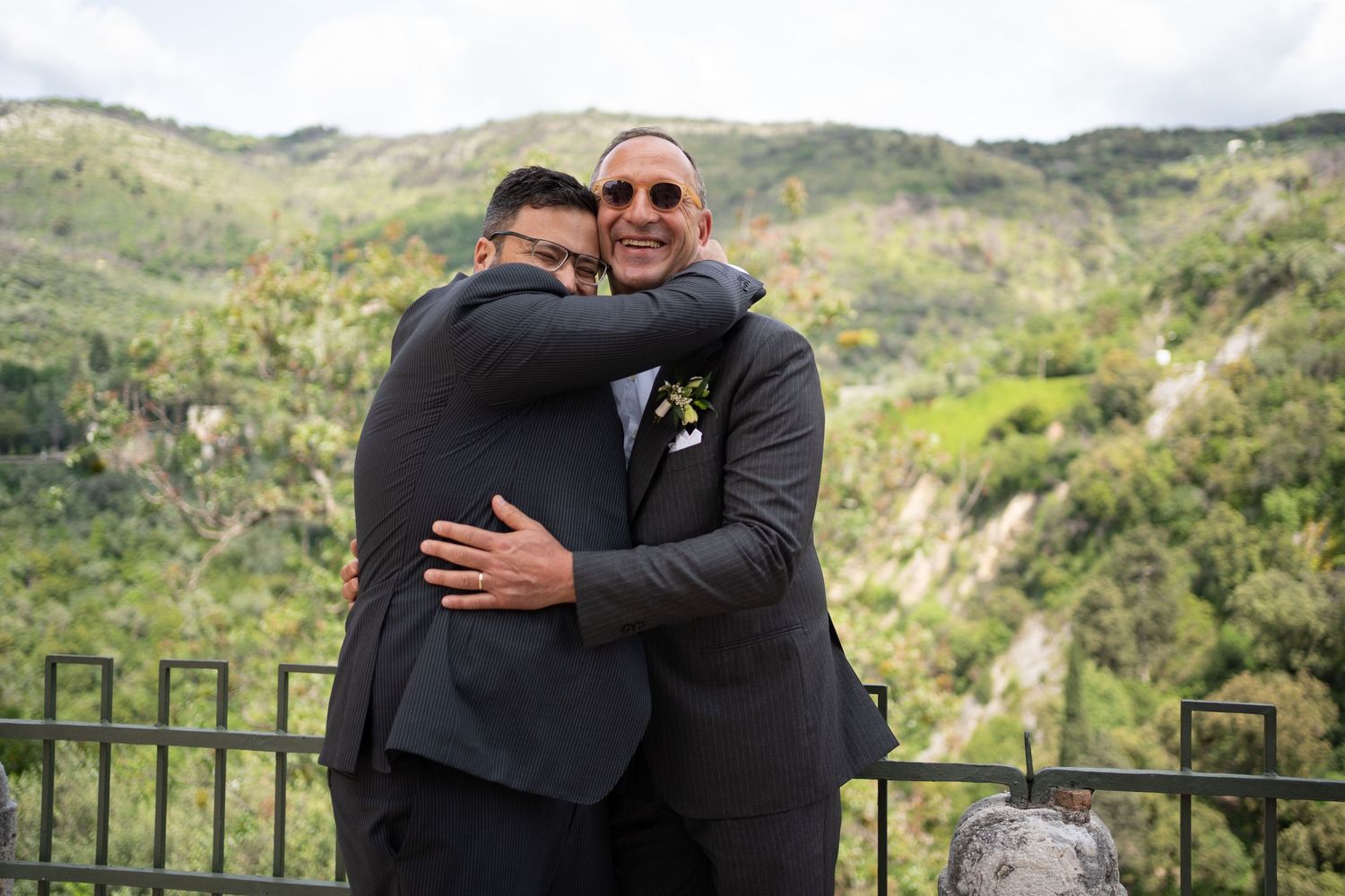 Two people embrace outdoors with scenic mountain landscapes and greenery in the background.