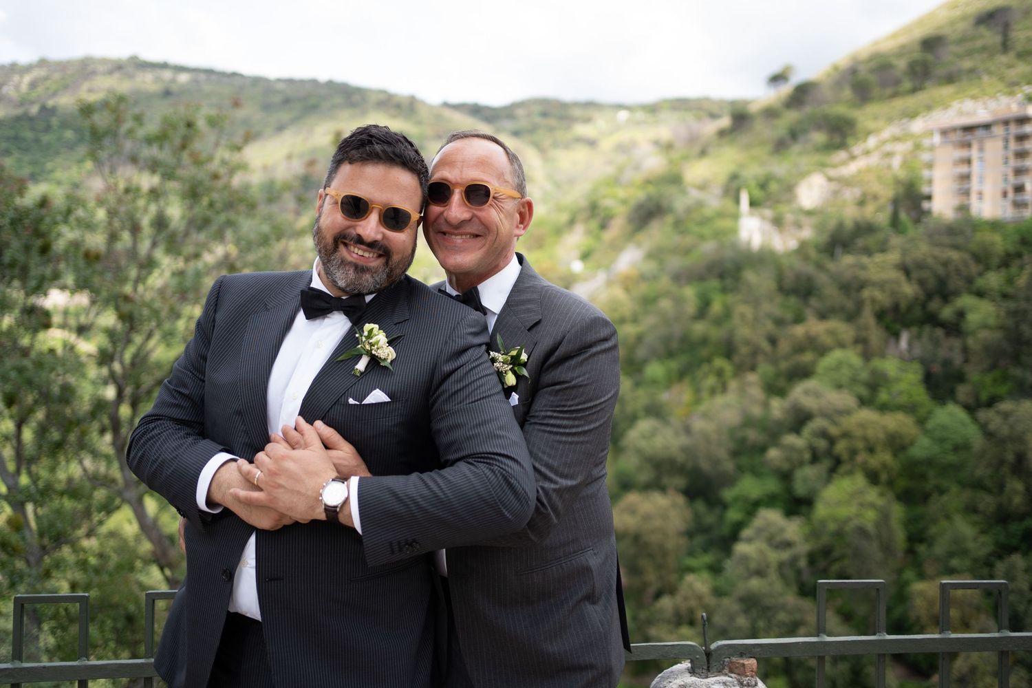 Two people in formal black suits embrace and smile while standing outdoors with green hills in the background.