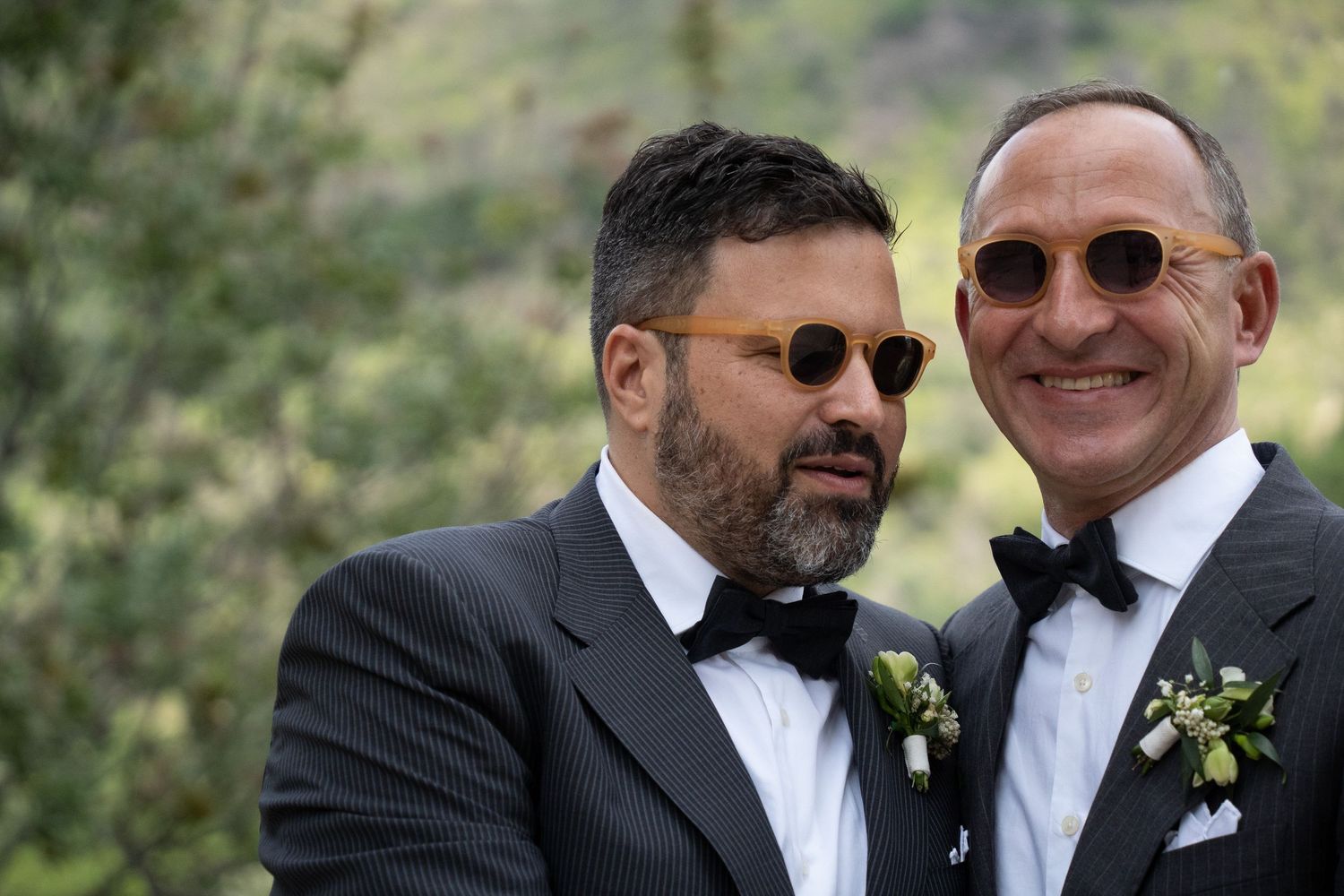 Two men in formal black tuxedos and bow ties smile while standing together at an outdoor event.