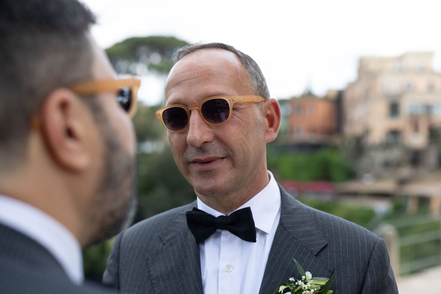 A wedding party poses together in formal attire with the groom wearing a black bowtie and sunglasses.