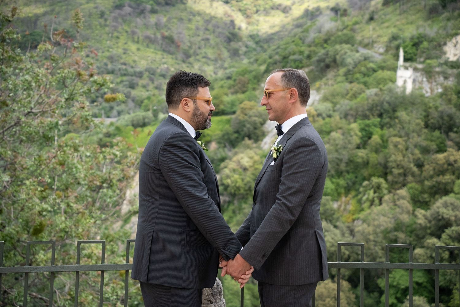 Two people in dark suits stand close together on a balcony with lush green mountains in the background.