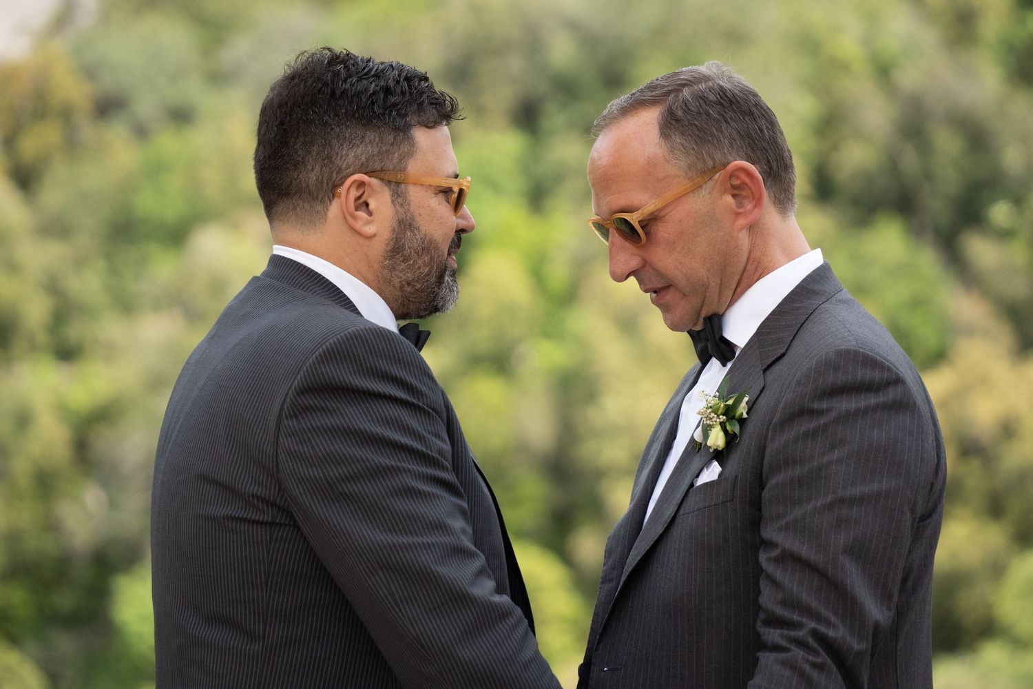 Two men in dark suits share an intimate moment during a wedding ceremony with greenery in the background.
