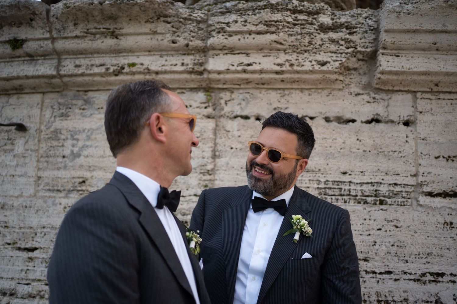 Two men in formal attire having a conversation against a white stone wall.