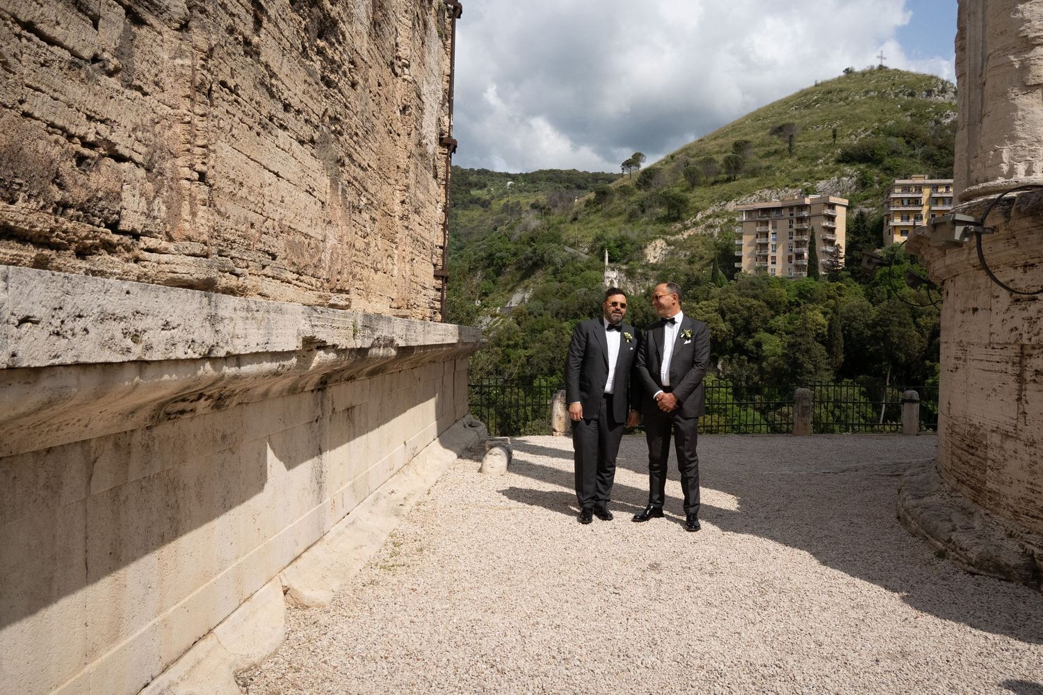 Two people in formal attire walk along a stone path with hillside buildings visible in the background.