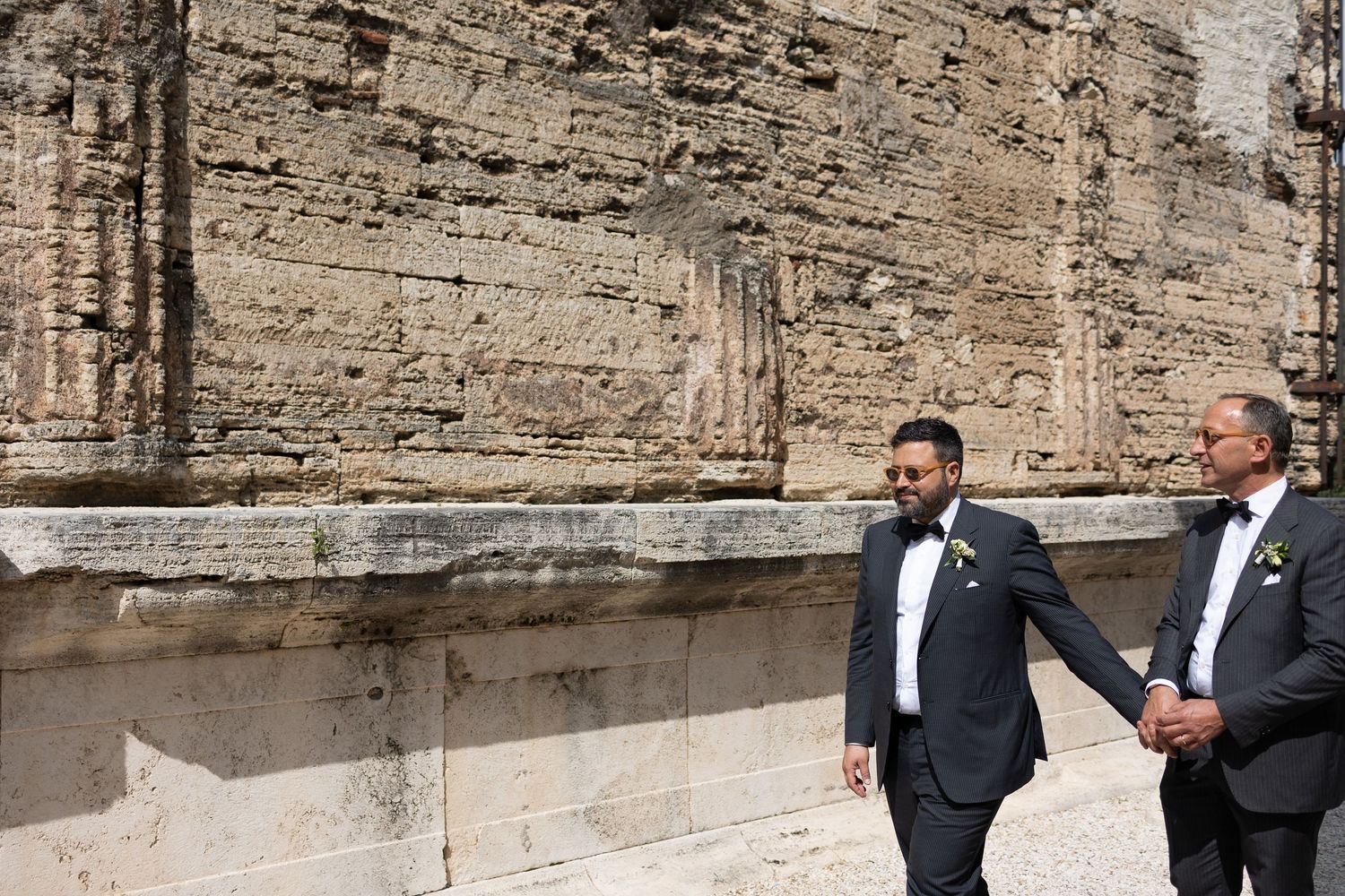 Two men in formal tuxedos stand together against a textured stone wall background.