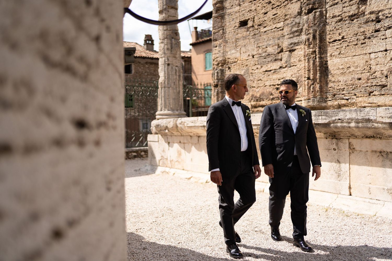 Two people in formal attire walk and chat together in front of a rustic stone wall on a sunny day.