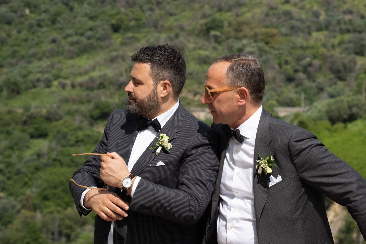 Two people in formal black suits share an embrace while standing on a green hillside.