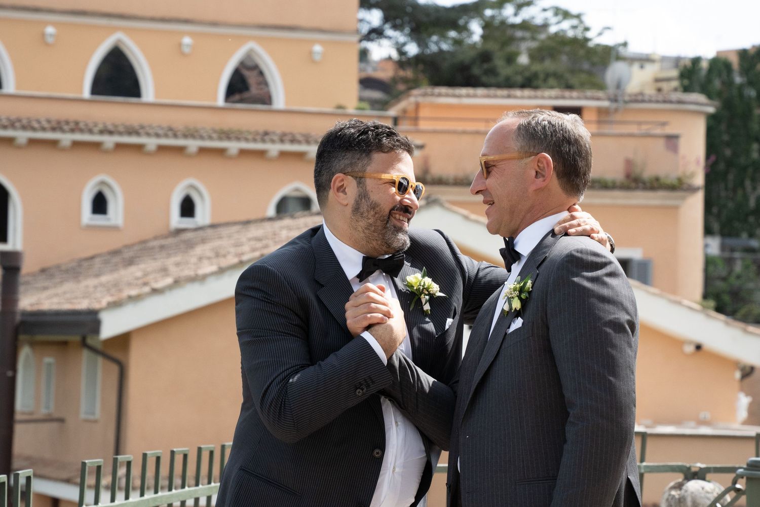 Two people in formal suits share an emotional moment at an outdoor wedding venue with Mediterranean-style architecture.