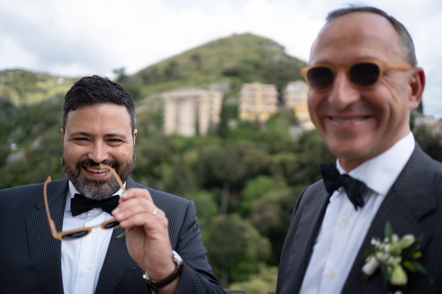 Two people in formal black tuxedos and bow ties smile together outdoors with a hillside building in the background.