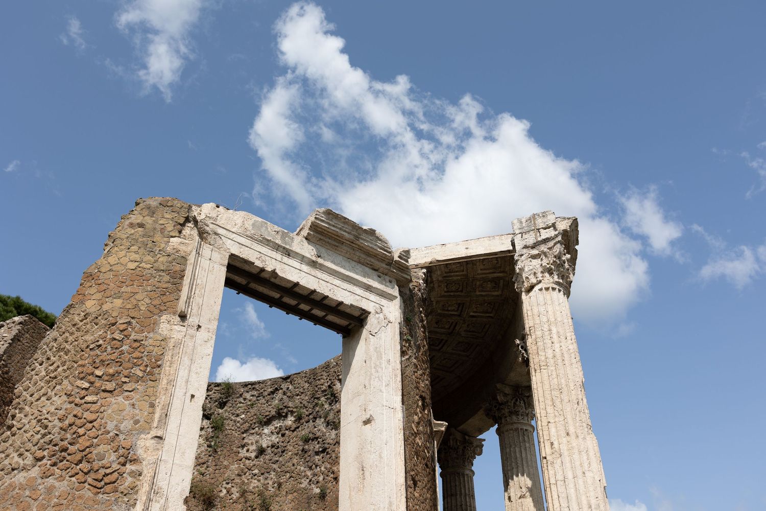 Ancient Roman temple ruins with white marble columns and stone walls against a bright blue sky with fluffy clouds.