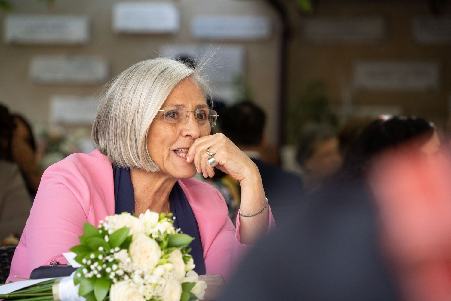 A person wearing a pink blazer sits at a table with white flowers and appears to be laughing or smiling.
