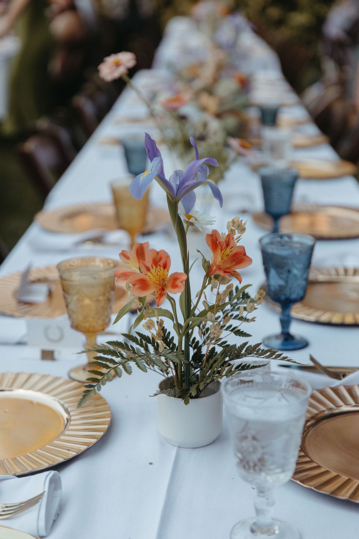 An elegant table setting with floral centerpieces featuring iris and lilies, blue and amber glasses, and gold-rimmed plates.