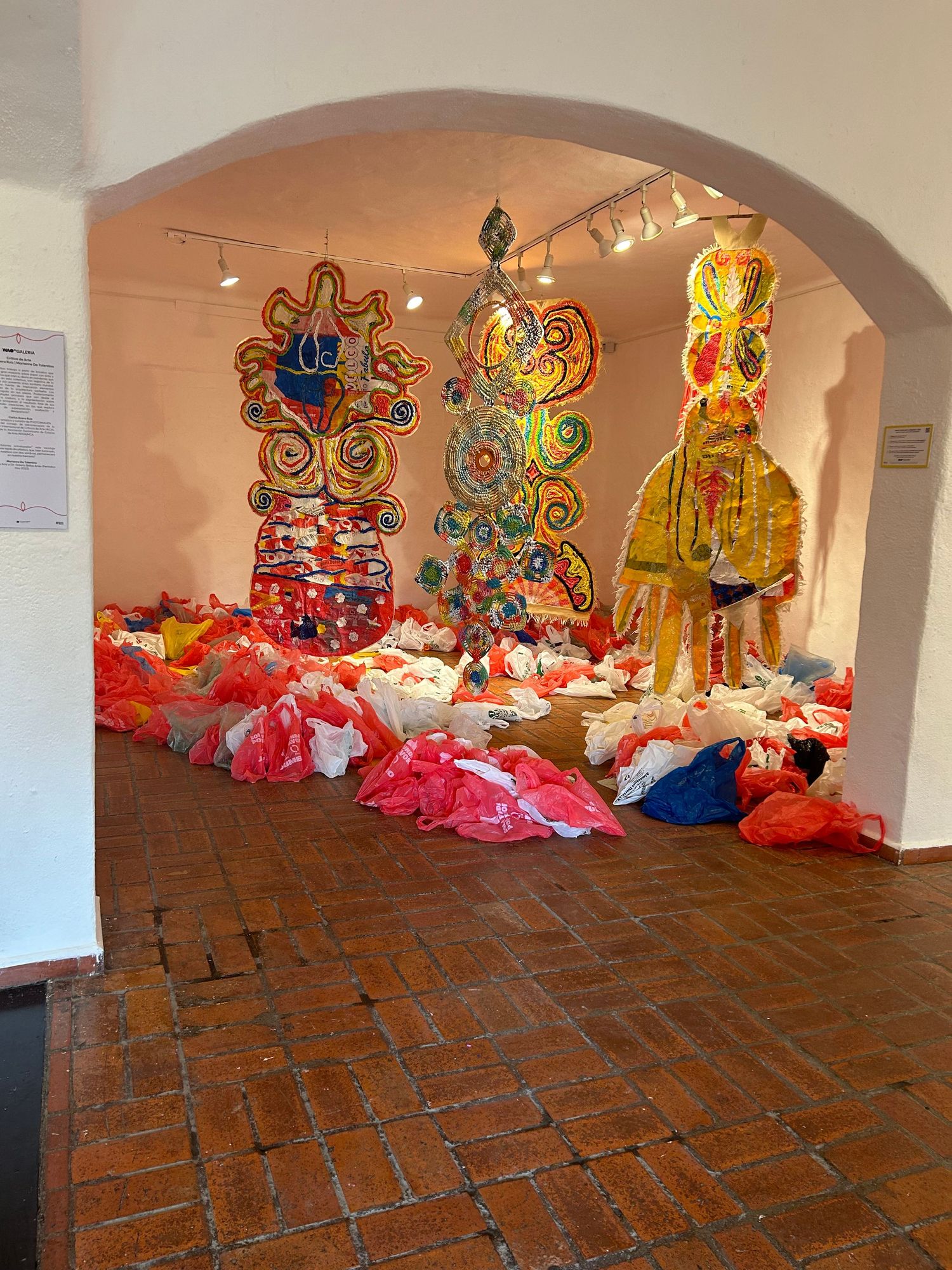 Art gallery space featuring tall colorful sculptures and red plastic bags arranged on brick tile floor.