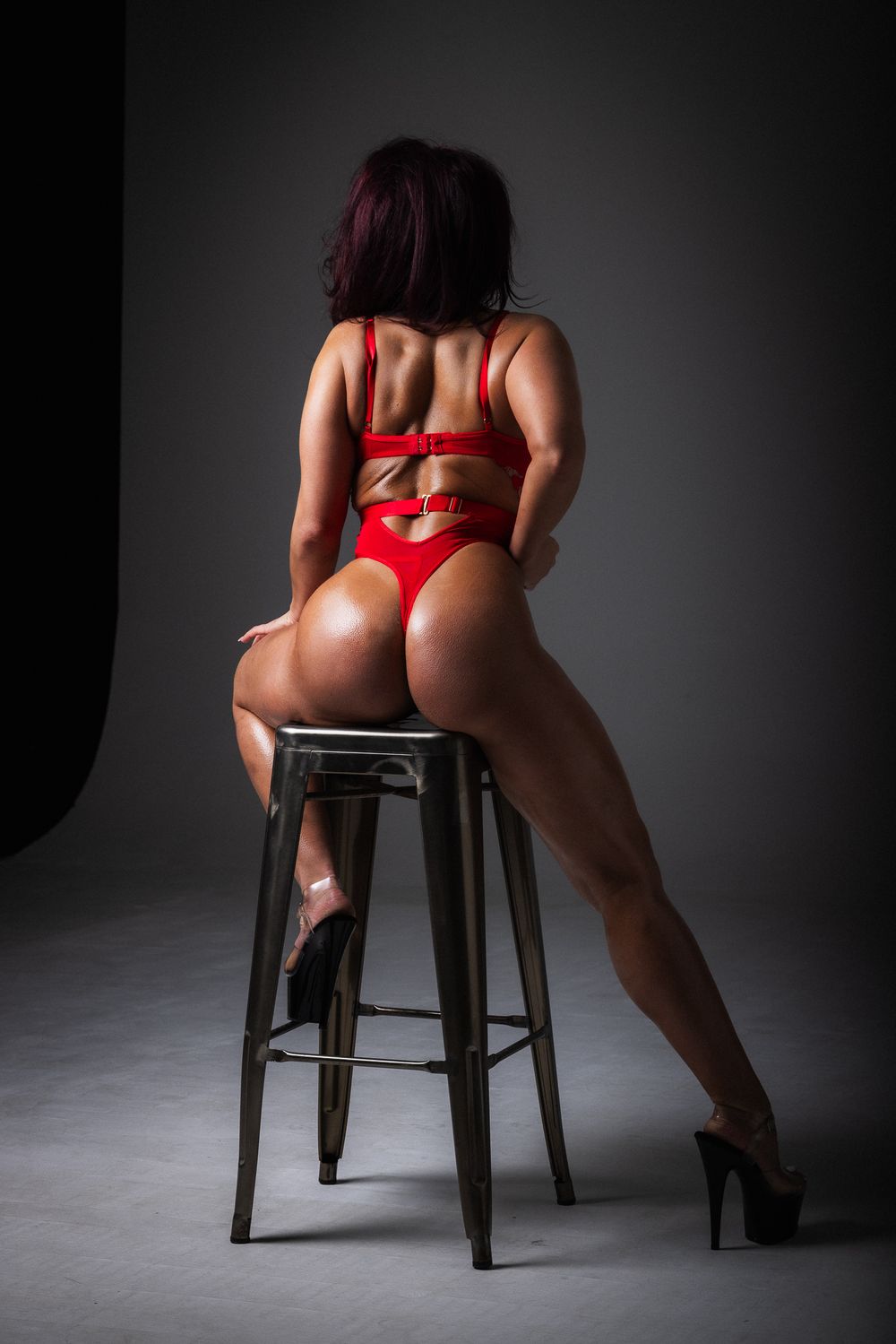 Back view of figure in red lingerie and heels posing on wooden barstool against dark background.