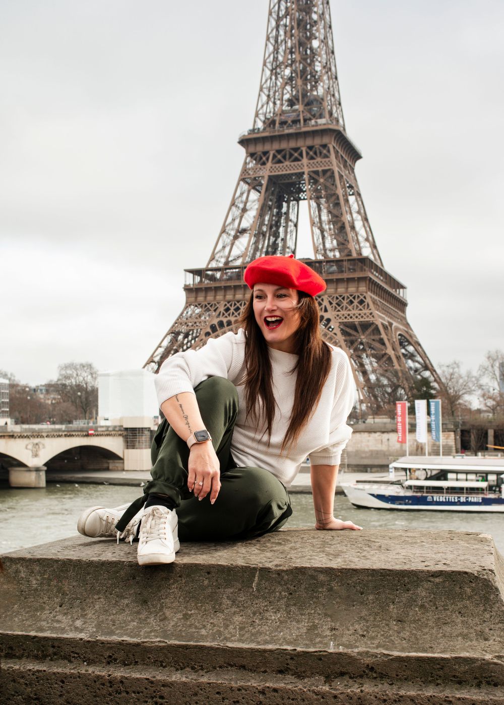 Tourist in red beret poses casually in front of the Eiffel Tower with Seine River and boats in background.