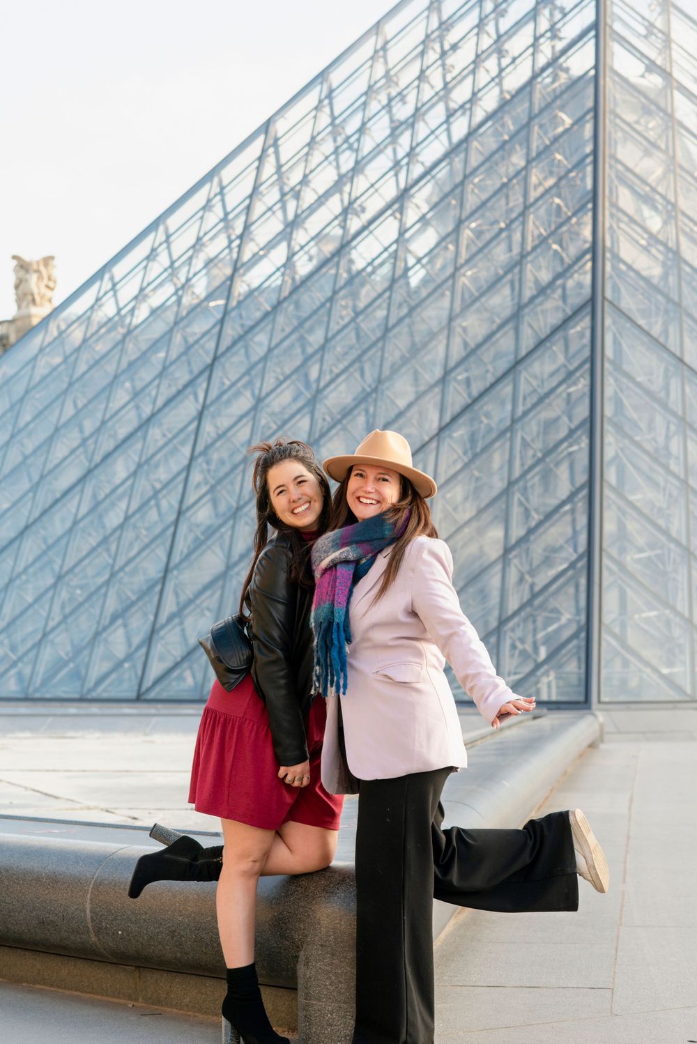 Two friends pose playfully in front of the glass pyramid at the Louvre Museum in Paris.
