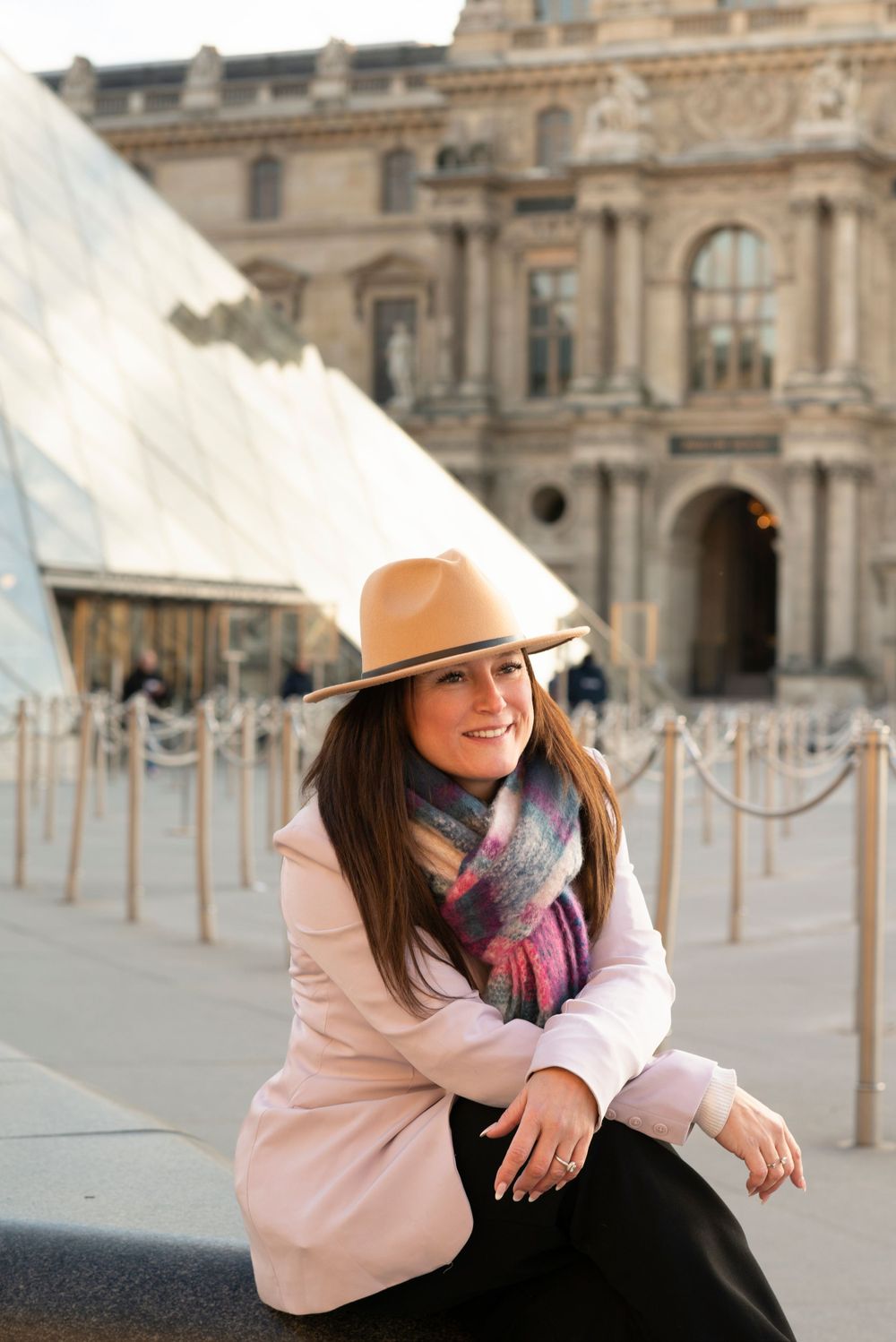 Stylish visitor in pink coat and tan hat poses near the Louvre Museum glass pyramid entrance.