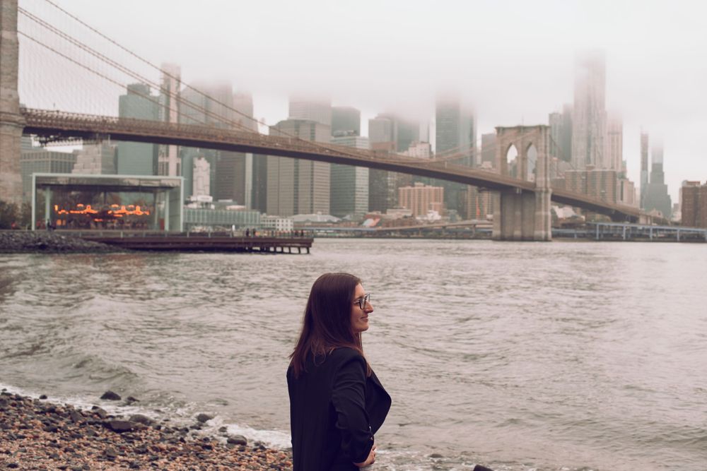 Person in dark coat looks out at Brooklyn Bridge and NYC skyline on foggy day.