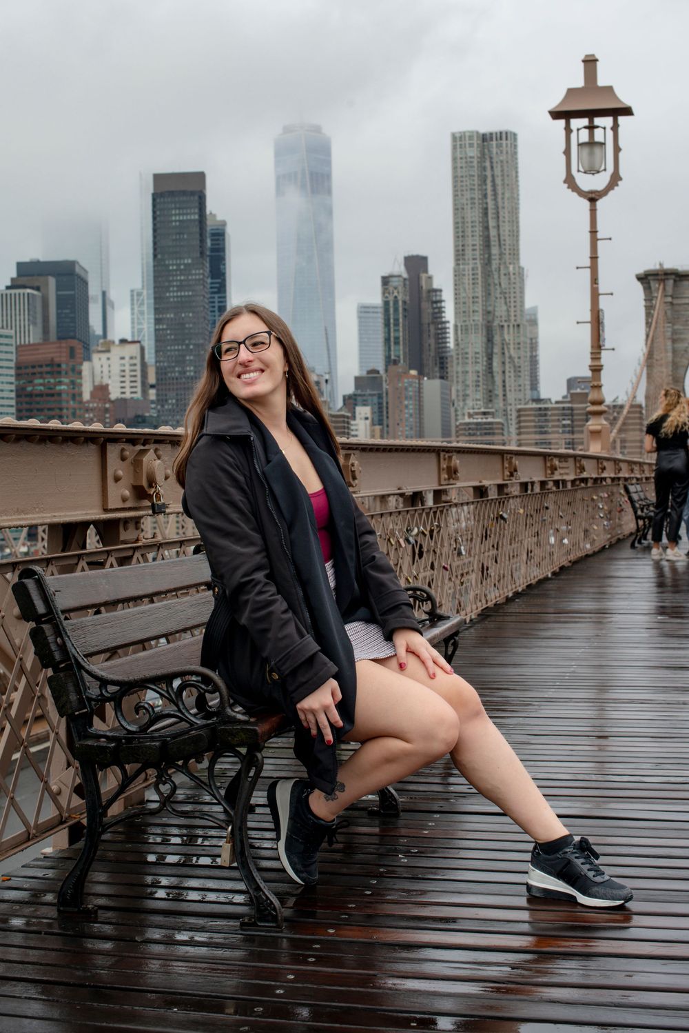 Tourist sits on bench on Brooklyn Bridge with Manhattan skyline visible through foggy weather.