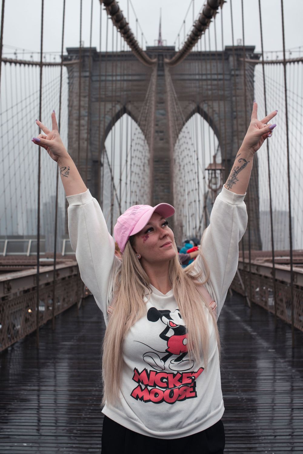Tourist in Mickey Mouse shirt makes peace signs on Brooklyn Bridge during foggy day.