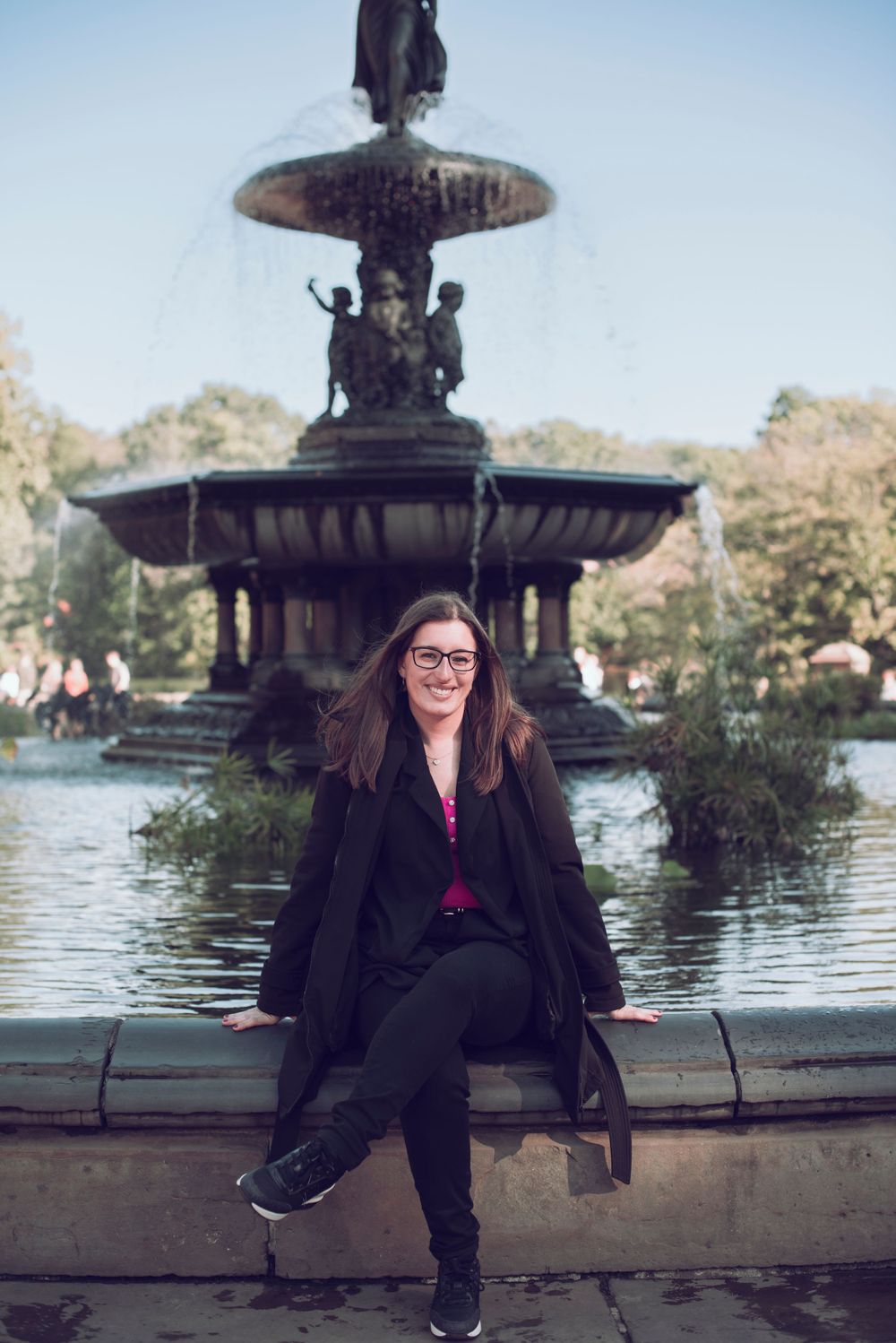 Tourist sits on edge of Bethesda Fountain in Central Park wearing black coat and glasses.
