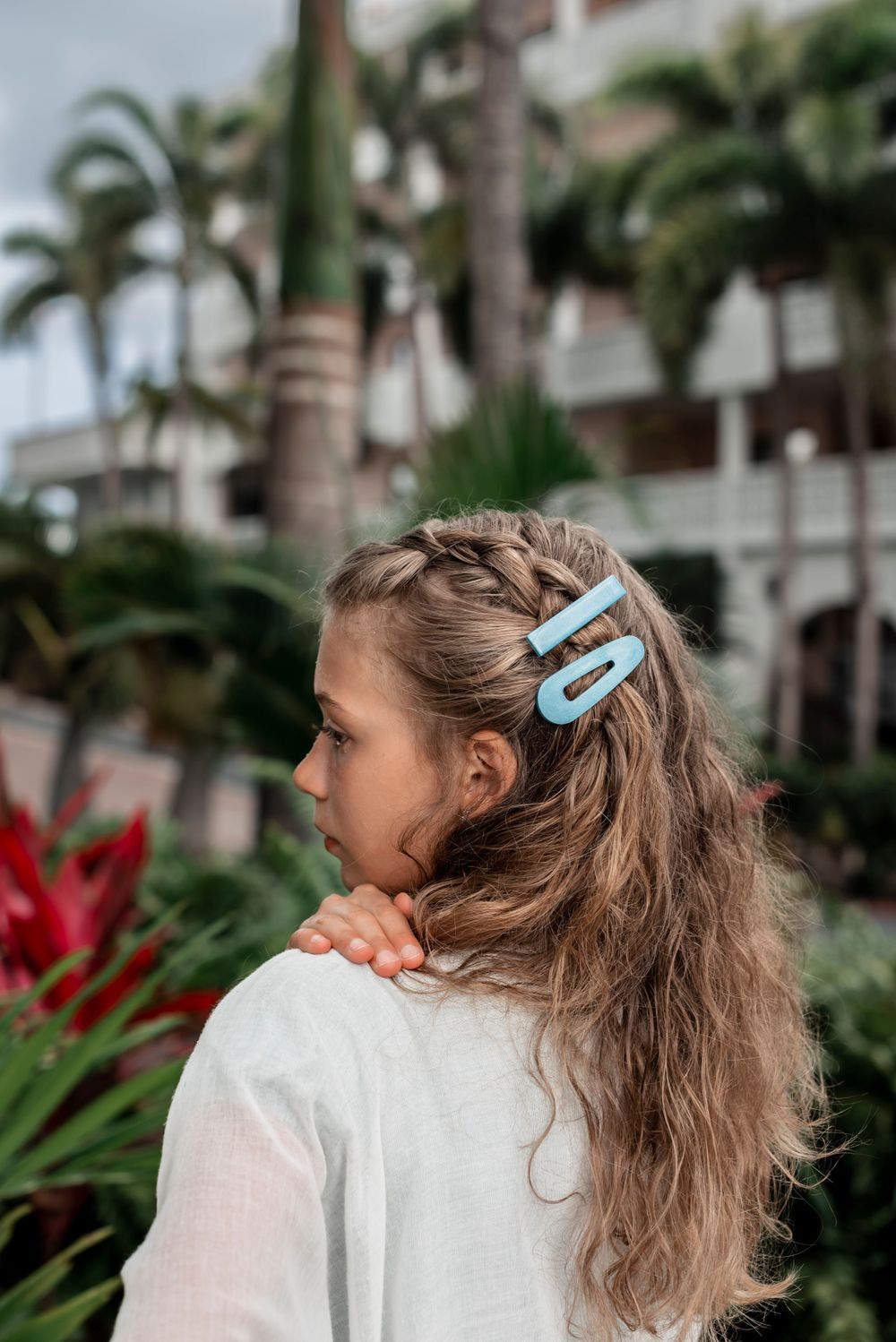 A person with long wavy hair wearing a white top and blue hair clips stands in a tropical garden with palm trees.