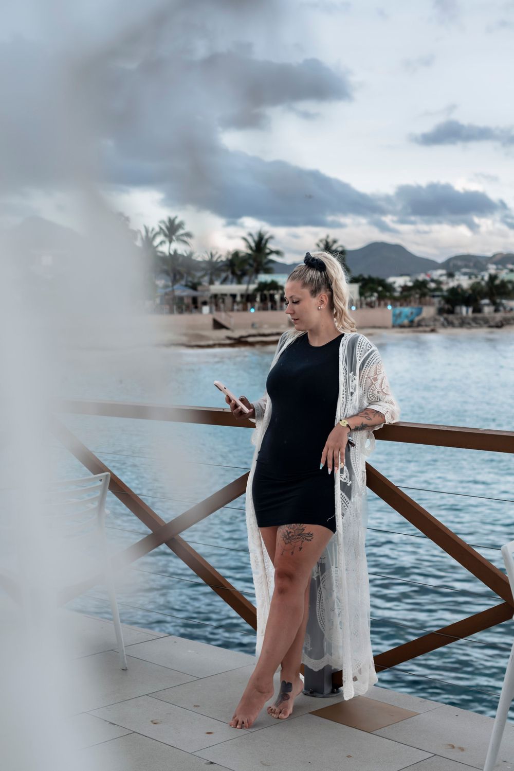 A woman in a black bodysuit and white kimono stands on a beachside deck with palm trees and mountains in the background.