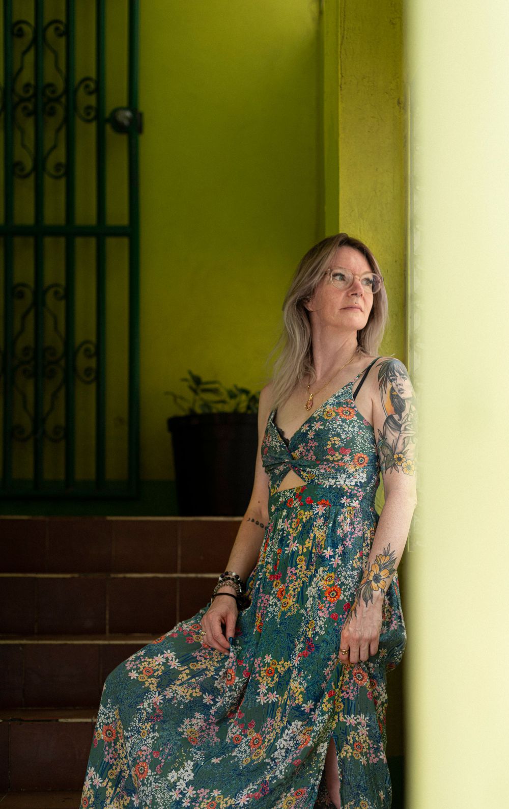 A woman in a teal floral maxi dress poses against a lime green wall with ornate ironwork detail.